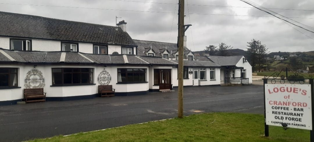 The whitewashed frontage of Logue's of Cranford under a grey Donegal sky, with benches at the windows and a roadside sign reading Logue's of Cranford: Coffee, Bar, Restaurant, Old Forge, Camper Van Parking.