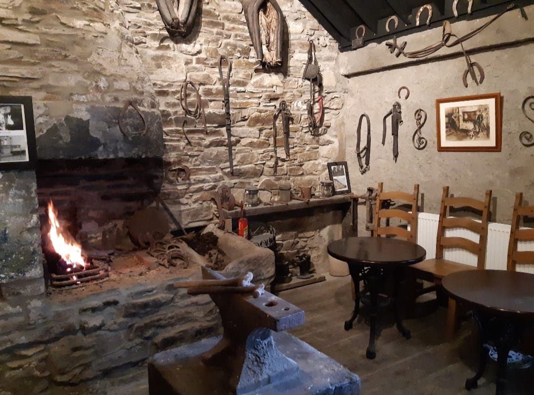 The Old Forge at Logue's: a lit fire in the original stone hearth, an anvil in the foreground, and horseshoes, tongs and blacksmith's tools hung on the stone walls beside small round tables.