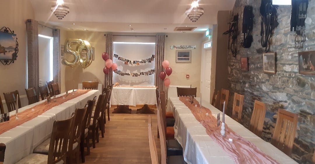 Logue's function room laid out for a 50th birthday: two long tables dressed in white linen with pink runners and candles, gold balloons, chandeliers, and an exposed stone wall hung with horse tack.